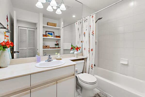 A modern bathroom with a white sink, toilet, and bathtub. There are shelves with decorative items and a mirror above the sink. - Elegant 2bd Townhouse with Loft, Lanais and views (Kihei)
