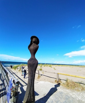 En skulptur af en kvinde på en strand med et træhegn og en klar, blå himmel.