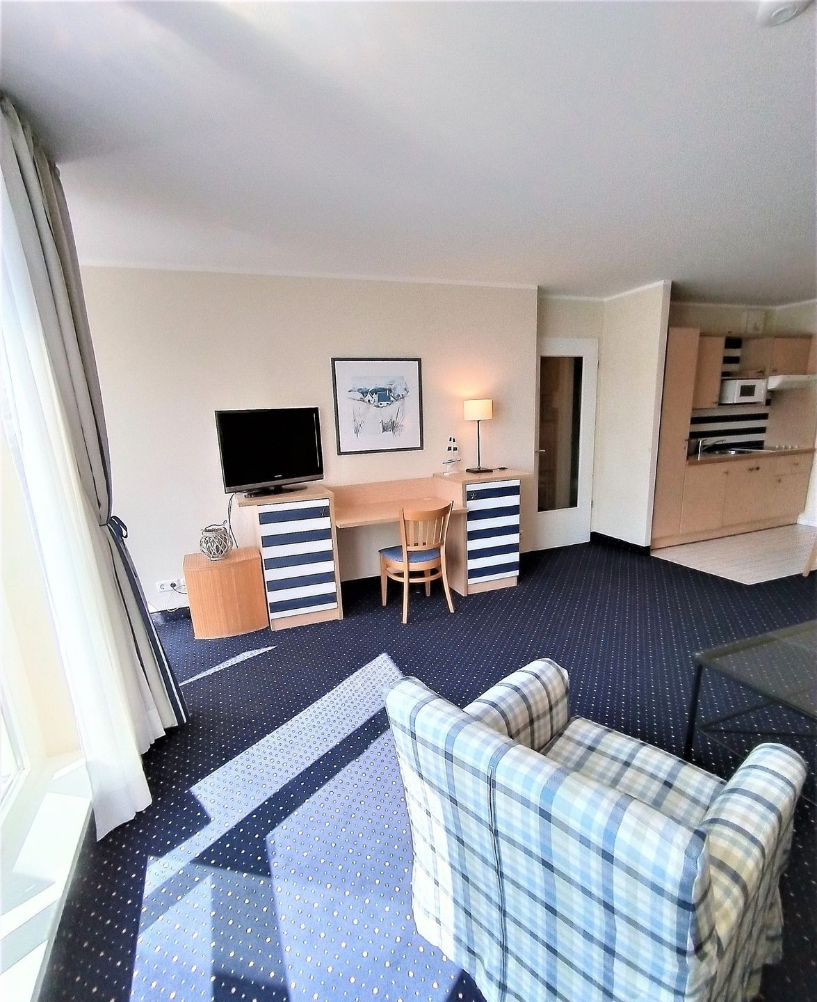 A hotel room with a bed, a desk with a chair, a television, a small table with a lamp, and a kitchenette.