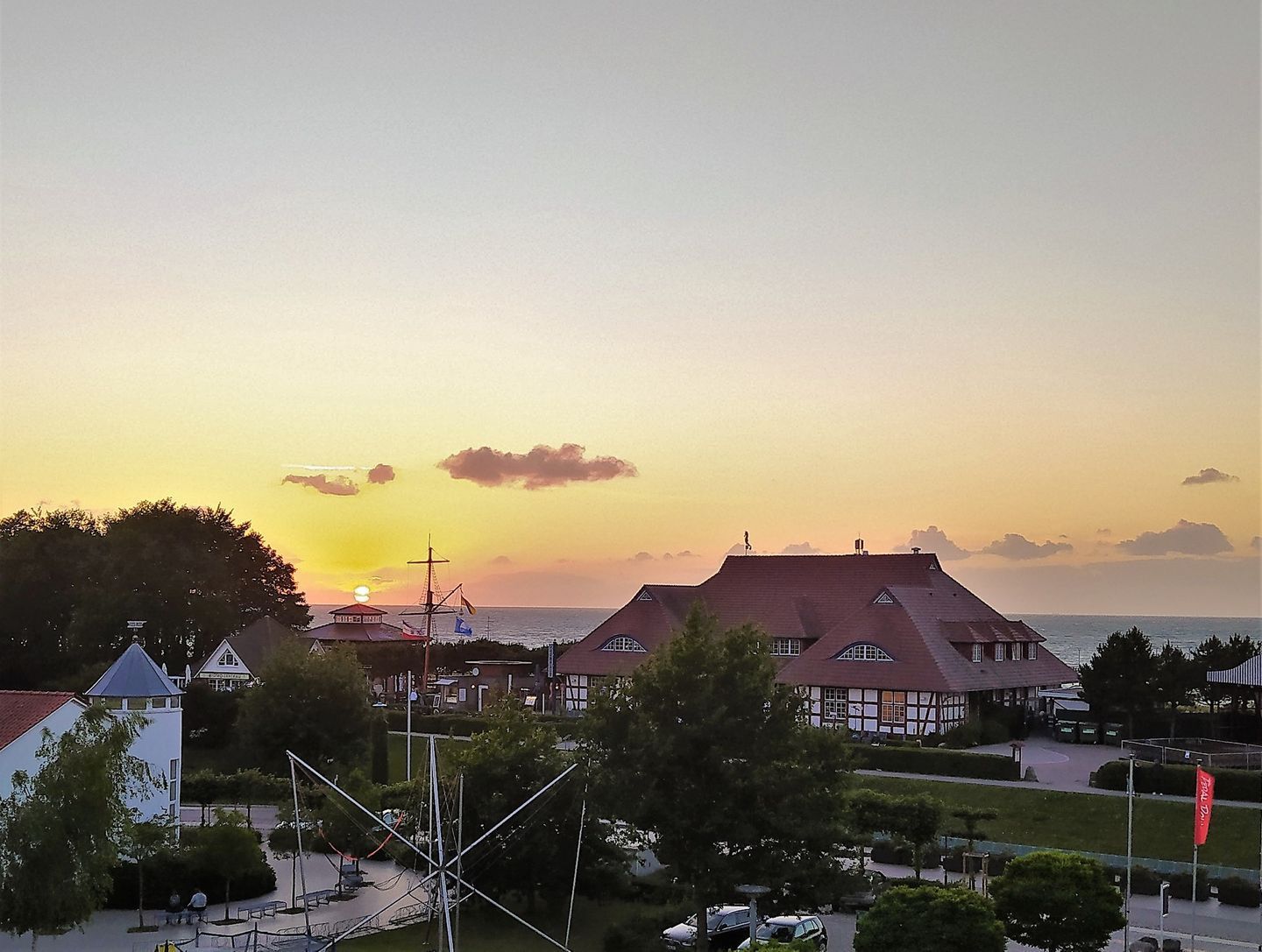 Une ville côtière au coucher du soleil, avec des bâtiments traditionnels, une église et un petit port où est amarré un voilier.