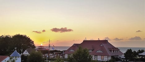 Une ville côtière au coucher du soleil, avec des bâtiments traditionnels, une église et un petit port où est amarré un voilier.