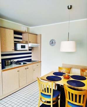 A compact kitchen with a microwave, stove, and sink, wooden cabinets, and a dining table with blue placemats and chairs. - "My time out" App. 202 - Private vacation apartment in a unique location directly on the (Zingst)