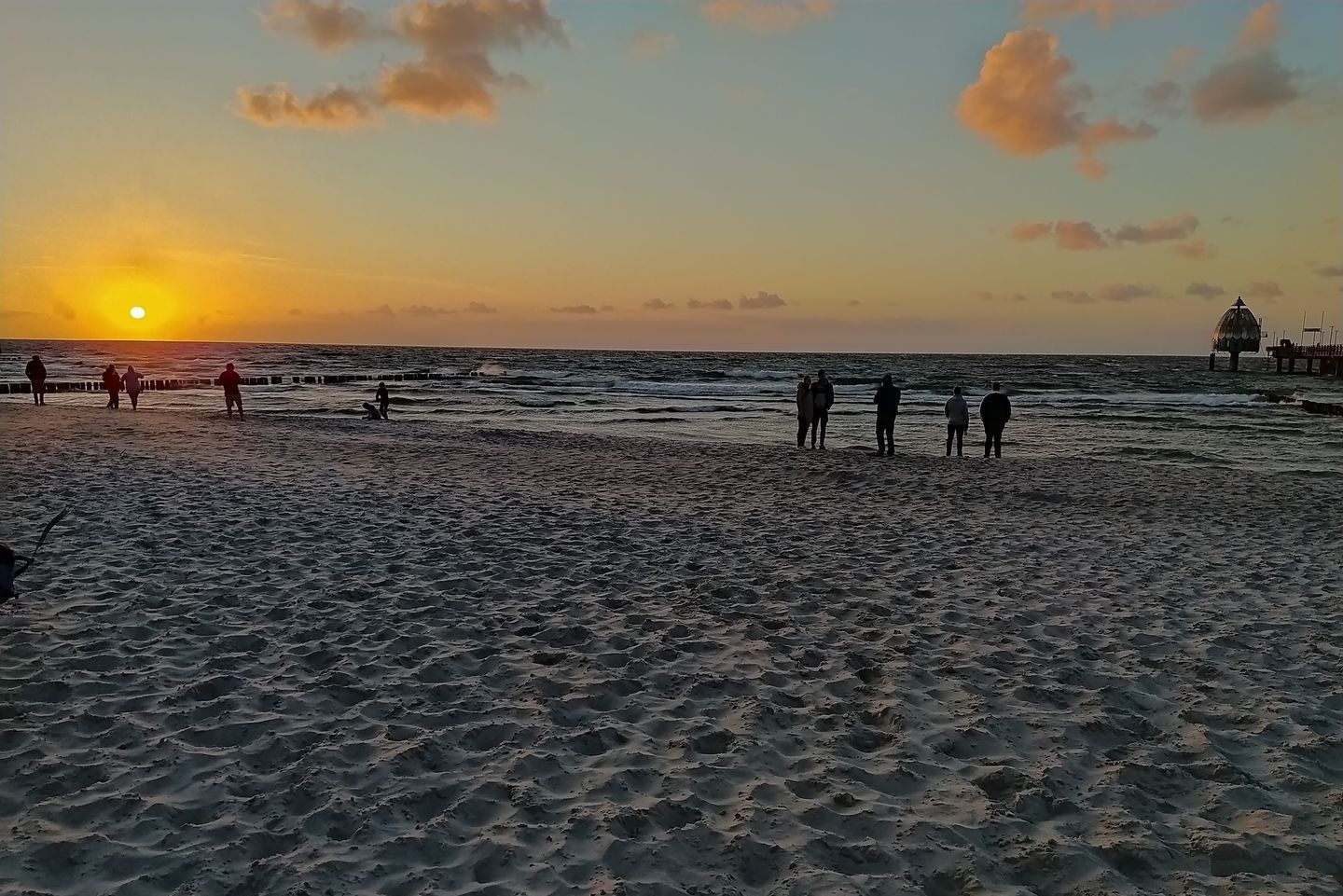 A beach with people walking and standing near the water during sunset.