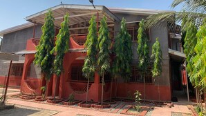 A building with a modern architectural design, featuring a red and grey color scheme, a covered outdoor area with a solar panel, and a garden with tall green plants.
