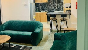 A modern living room with green sofas and a small wooden table. A kitchen with a black and white backsplash and wooden cabinets is visible in the background.