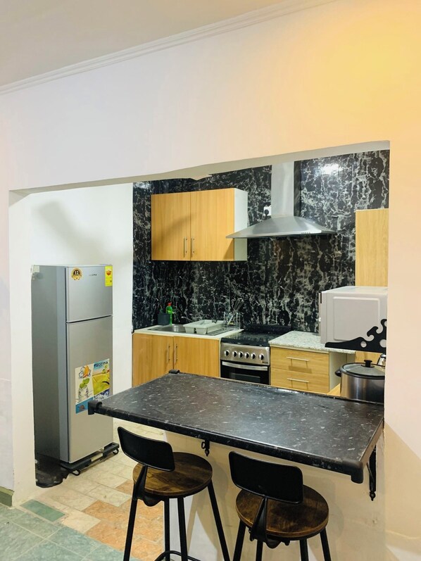 Cocina compacta con salpicadero de mármol negro, armarios de madera, estufa de acero inoxidable y una pequeña isla con dos taburetes.