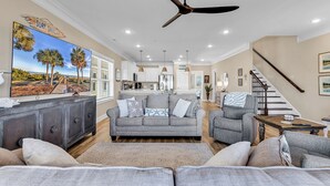 A spacious living room with a large flat-screen TV, a ceiling fan, and a kitchen in the background. - Flew The Coop-Second Row Beach House Community (Garden City Beach)