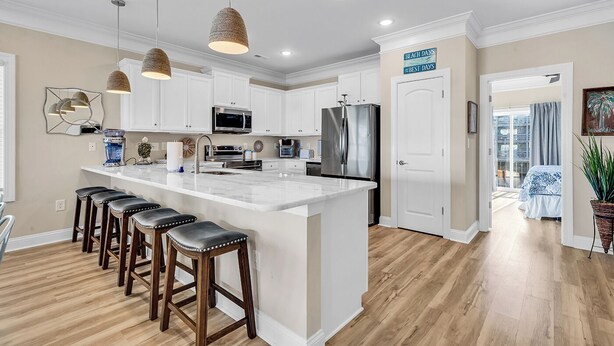A modern kitchen with white cabinets, stainless steel appliances, and a central island with bar stools. - Flew The Coop-Second Row Beach House Community (Garden City Beach)