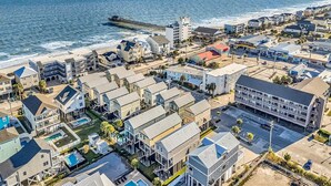 A coastal residential area with a pier extending into the ocean, featuring a mix of single and multi-story houses, a parking lot, and a small commercial building. - Flew The Coop-Second Row Beach House Community (Garden City Beach)