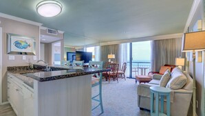 Une chambre d’hôtel avec un coin cuisine, une table à manger avec des chaises, un canapé et un balcon avec des rideaux.