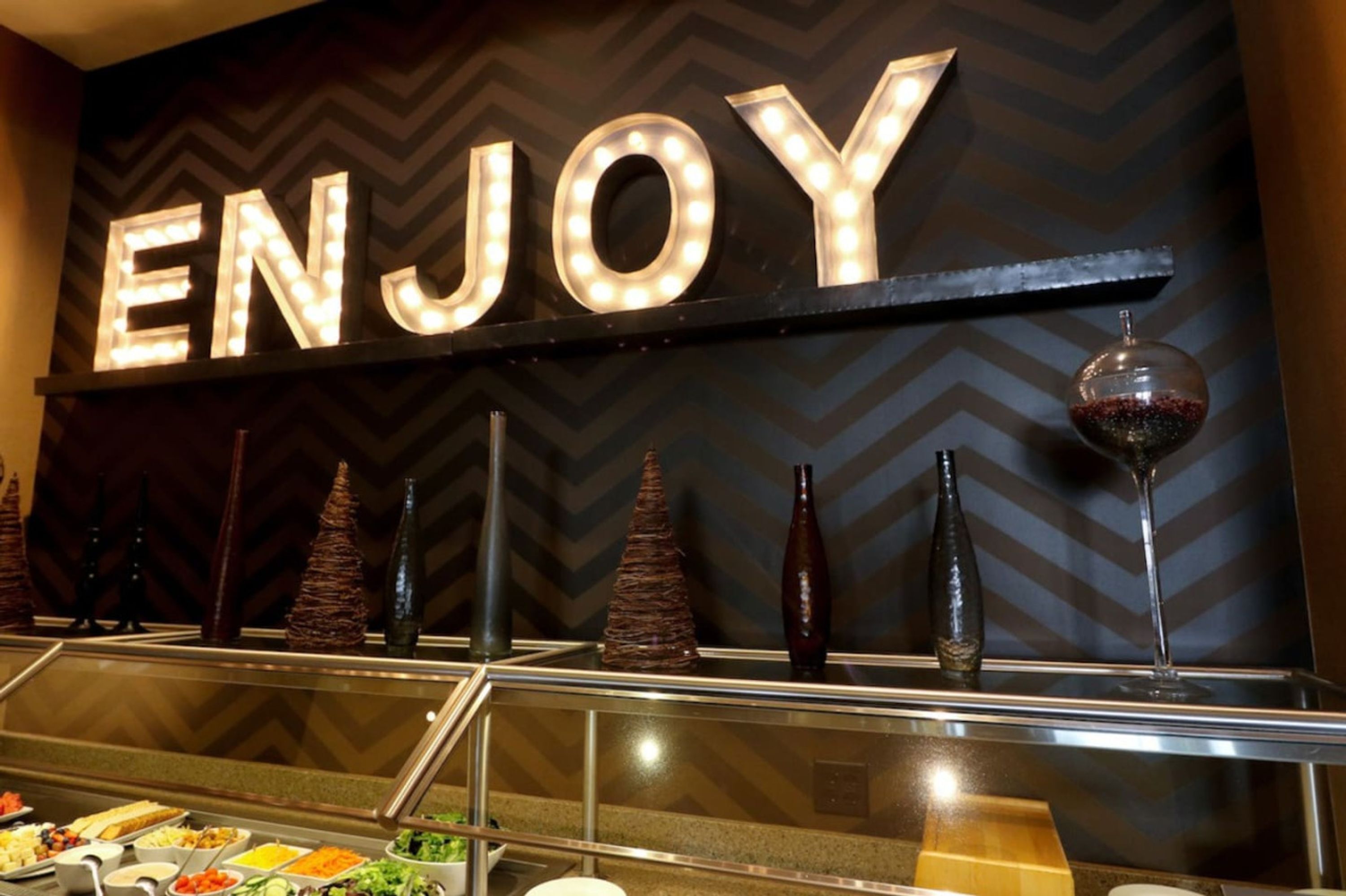 Un'area buffet con un cartello che recita “ENJOY” e diversi cibi e bottiglie decorative.