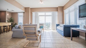 A spacious living area with a ceiling fan, a large window with blinds, a blue sofa, a wooden chair, and a kitchen counter with stools.