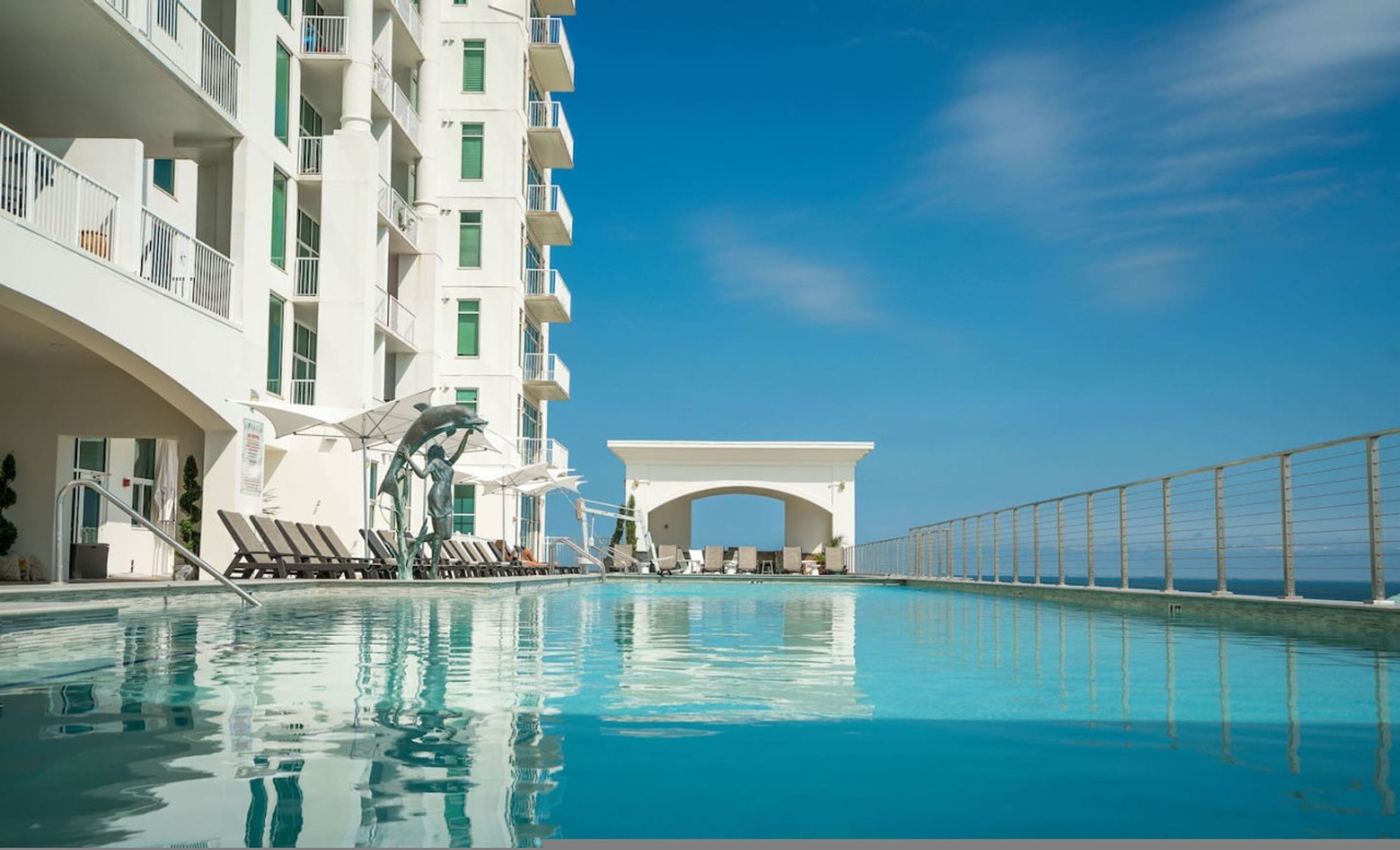 Kirkkaan sinisen taivaan alla oleva uima-allas, valkoinen rakennus parvekkeineen ja koristeellinen delfiinipatsas.