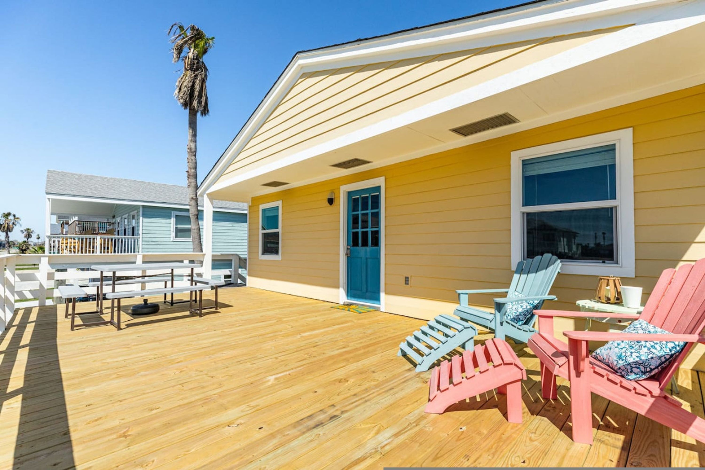 A wooden deck with outdoor seating, a palm tree, and a yellow building with a blue door and windows.