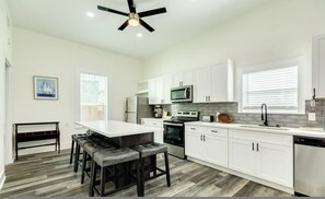 A modern kitchen with white cabinets, a central island with stools, a ceiling fan, and a large window with blinds. - Adventure Awaits Close to the Beach (Galveston)