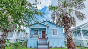 A light blue two-story house with a front porch, a wooden staircase, and a palm tree.