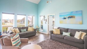 A spacious living room with two grey sofas, a coffee table, and a ceiling fan. Large windows provide a view of the outside. - Great bay Views and a Pool in Isla Del Sol (Galveston)