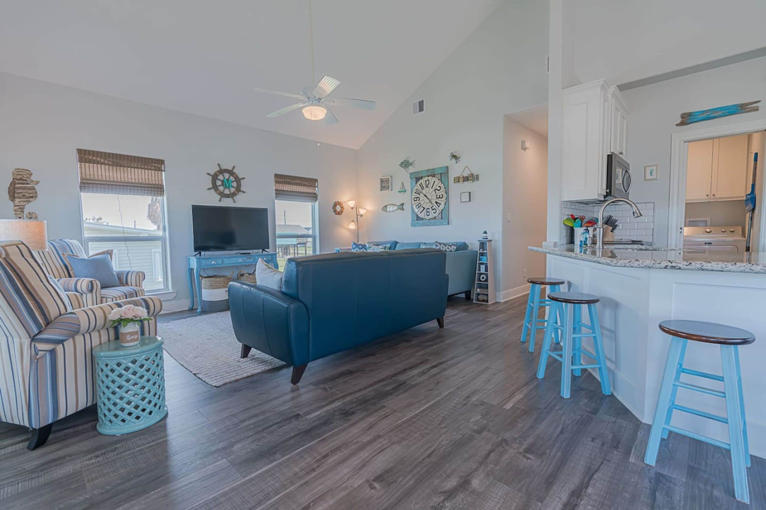 A spacious living room with a blue sofa, striped armchair, and a TV. There is a kitchen area with a bar and stools.
