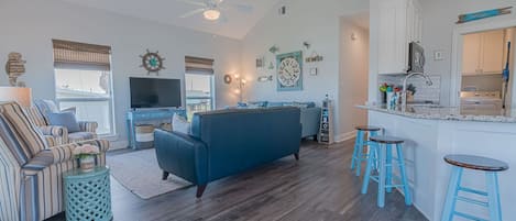 A spacious living room with a blue sofa, striped armchair, and a TV. There is a kitchen area with a bar and stools.