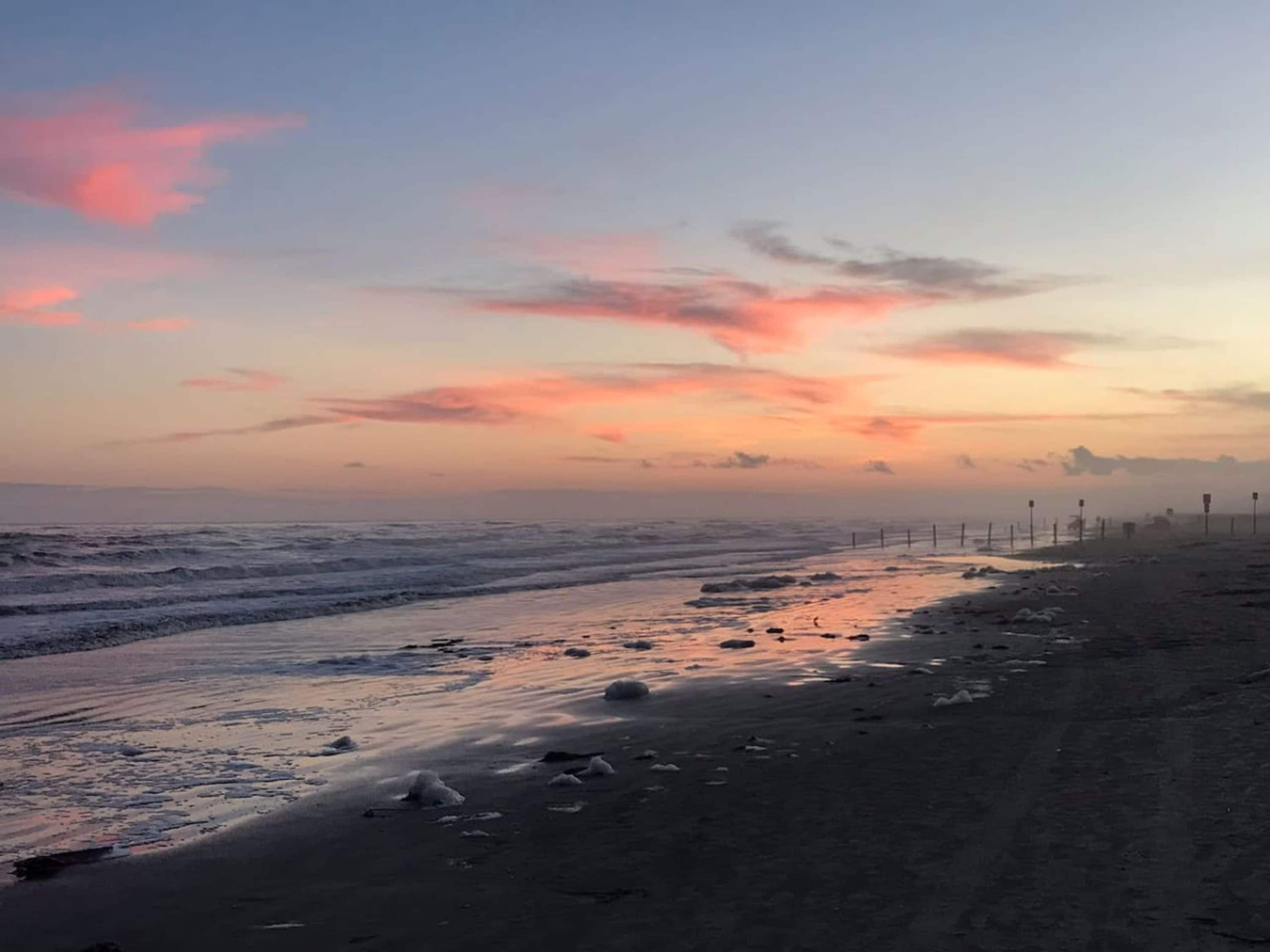 En strand med ett staket längs strandkanten, vågor som bryter mot sanden och en solnedgång i bakgrunden.