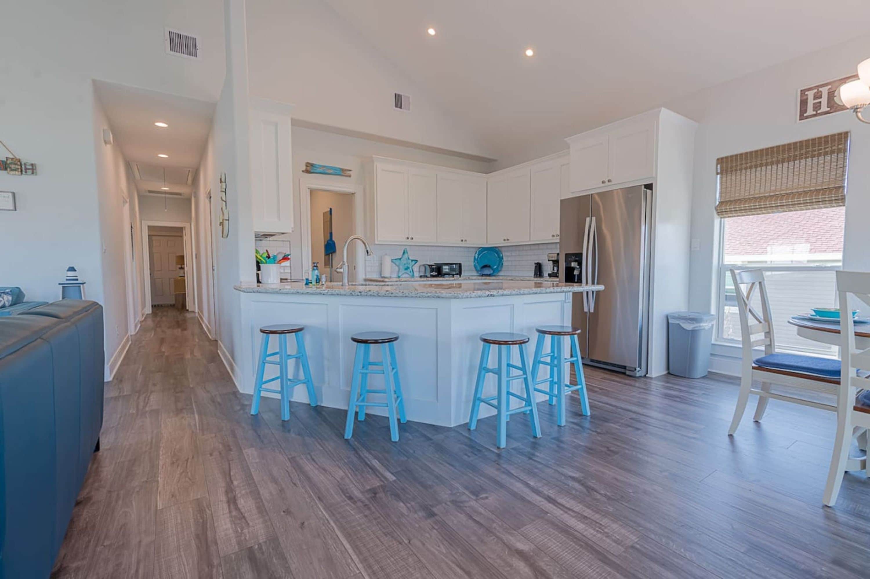 Una cocina moderna con armarios blancos, una isla central con taburetes, un refrigerador de acero inoxidable y un comedor con mesa y sillas.