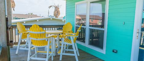 Un espacio al aire libre techado con un muro turquesa, una terraza de madera y sillones Adirondack amarillos alrededor de una pequeña mesa redonda.