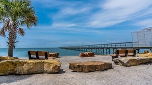 A beach with a palm tree, benches, and a pier extending into the water. - The Coast is Clear Great Home By Beach Bay (Galveston)