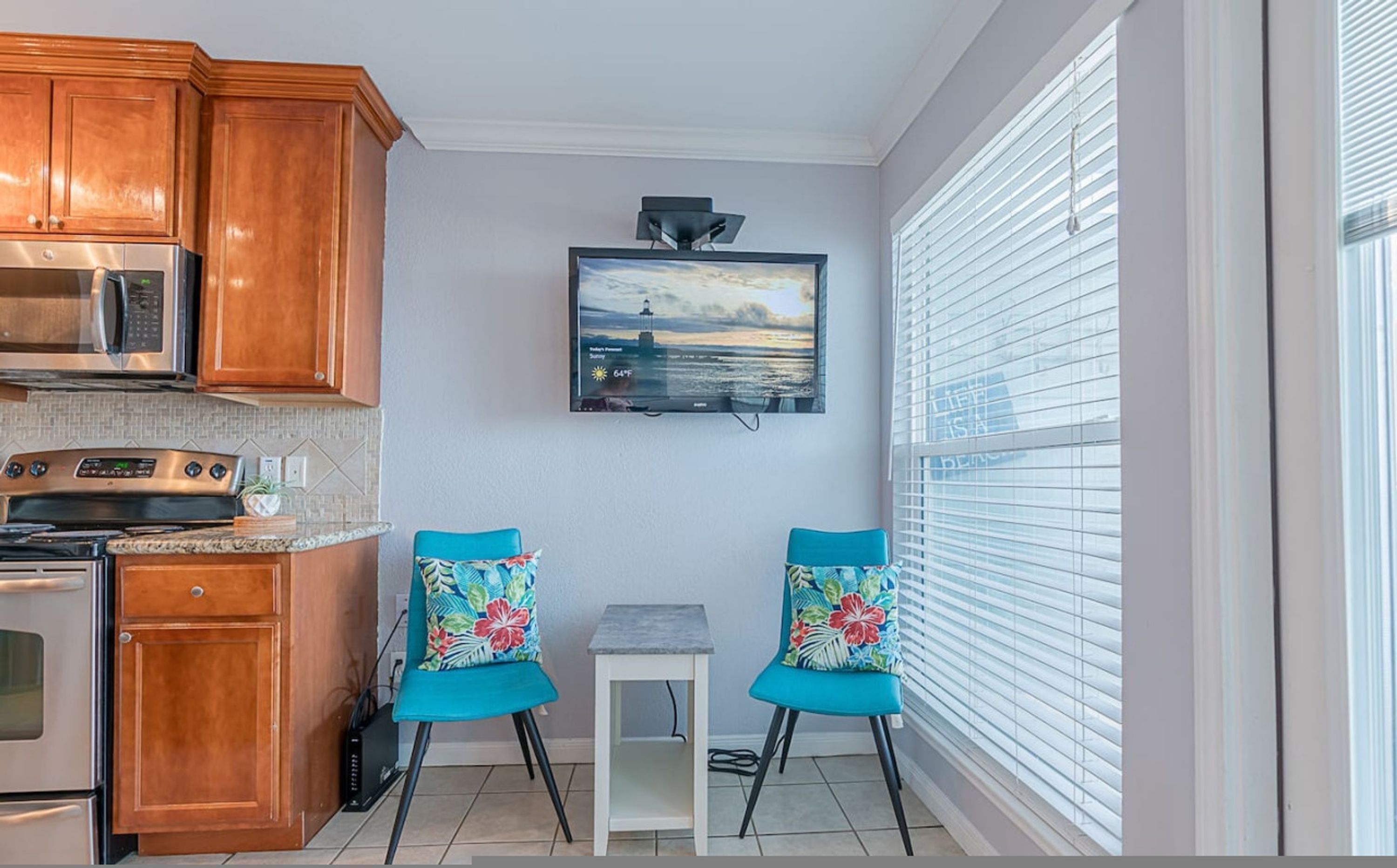 A compact kitchen with wooden cabinets, a microwave, an oven, and a small table with two chairs. A painting of a lighthouse is on the wall, and there is a window with blinds.