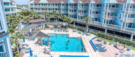 A resort with a central swimming pool surrounded by lounge chairs and umbrellas, multiple balconies, and a clear view of the ocean.