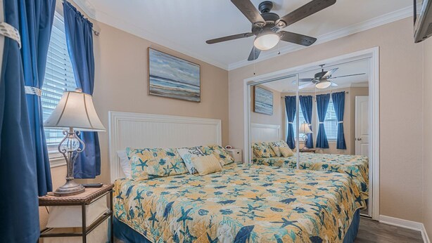A bedroom with a double bed, a ceiling fan, a nightstand with a lamp, and a window with curtains. - Get Beach Dazed in Galveston (Galveston)