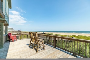 Une terrasse en bois avec deux fauteuils à bascule et une chaise rouge, donnant sur une plage bordée d’une clôture en bois et sous un ciel dégagé.