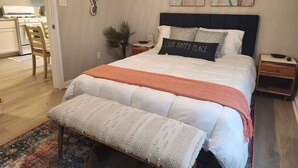 A bedroom with a bed, a bench, a nightstand, a wall-mounted lamp, and a kitchen visible through an open door. - Vacation in Style at the Beach (Galveston)