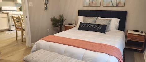 A bedroom with a bed, a bench, a nightstand, a wall-mounted lamp, and a kitchen visible through an open door.