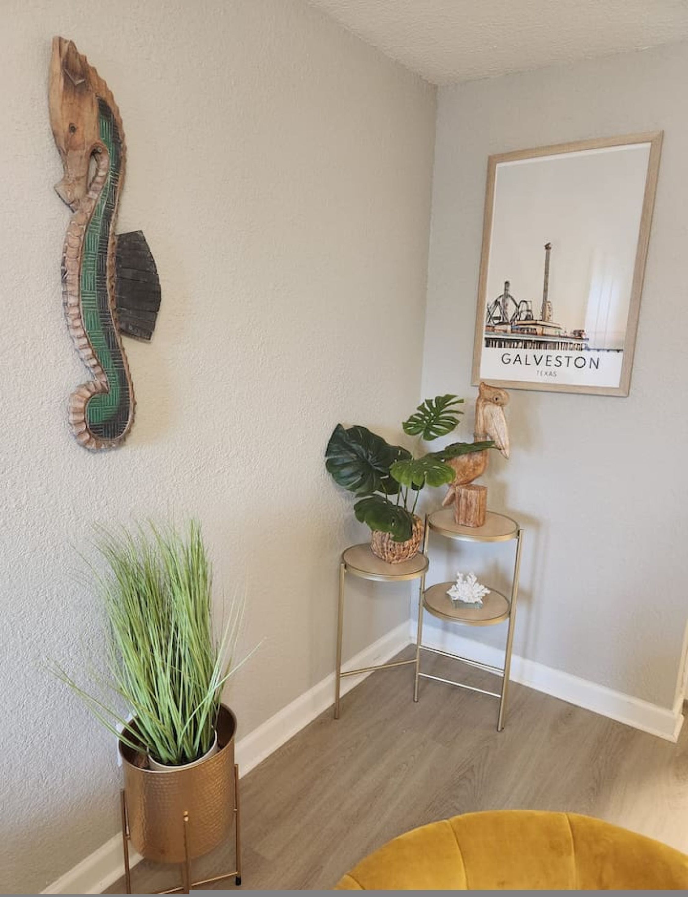 Ein Blick in einen Raum mit einer Holzdekoration an der Wand, einem gerahmten Bild von Galveston, Texas, einer kleinen Pflanze im Goldtopf und einem gelben Sessel.