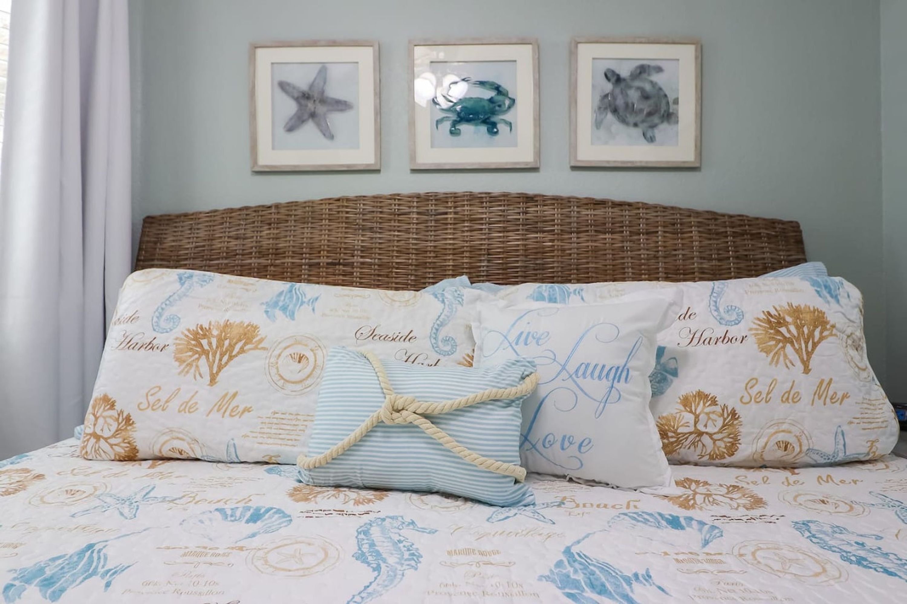 Un letto con testiera intrecciata, numerosi cuscini con decori marini e quadri appesi al muro.