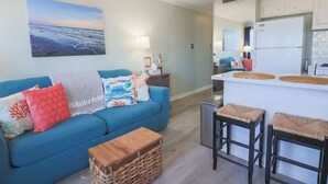 A living room with a blue sofa, a small wooden table, and a kitchen area with a refrigerator and bar stools.