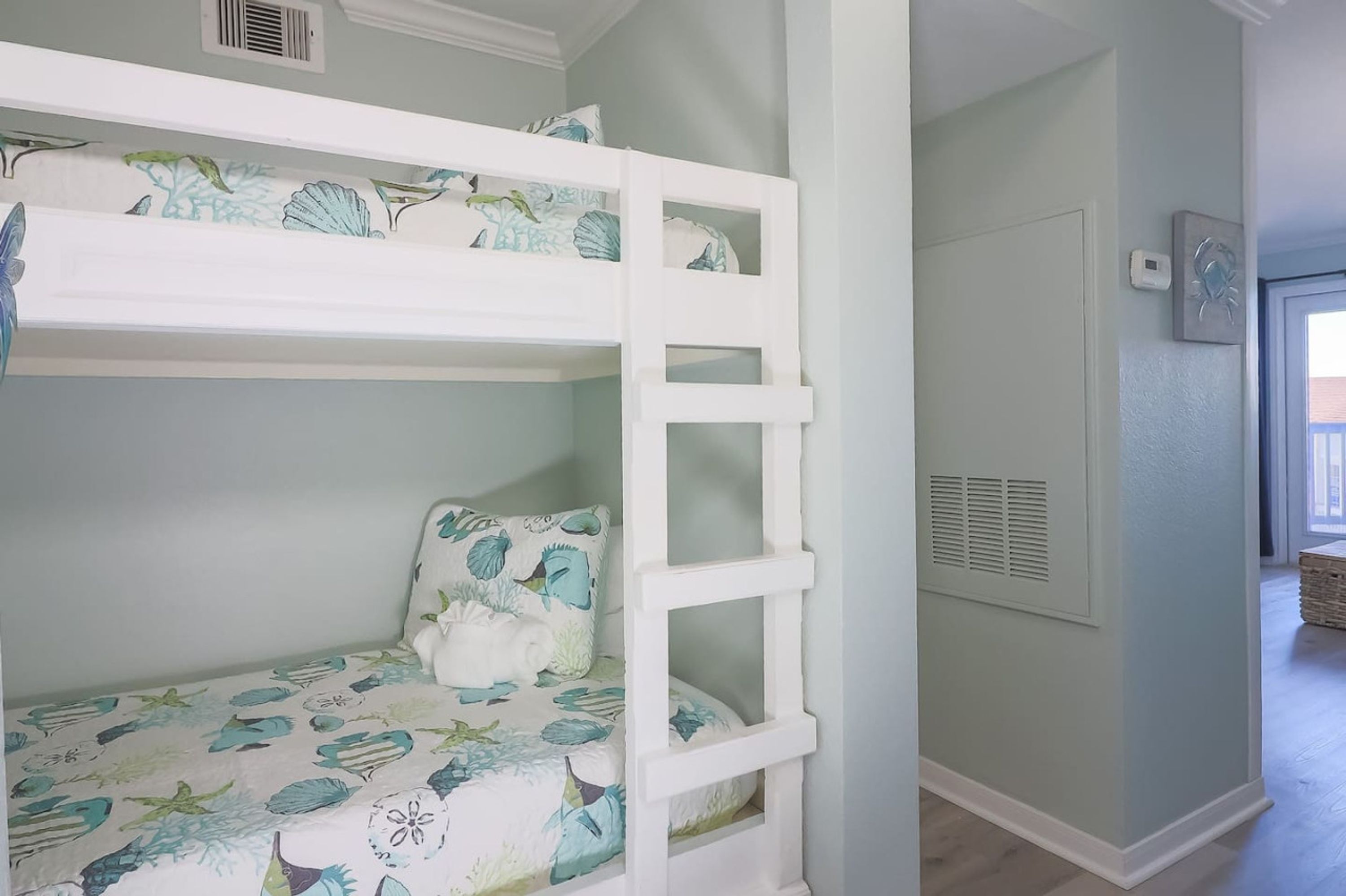Uma cama beliche com colcha florida, uma parede branca com um respiradouro, e uma porta que dá para outro quarto.