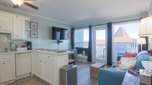 A modern kitchen with white cabinets, a coffee bar, and a built-in oven. There is a dining area with a blue sofa and a table. The room has large windows with a view of a building outside.