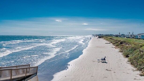 Ein Strand mit einem Holzsteg, einem Stuhl und einer Reihe von Wohnmobilen rechts.