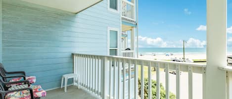 A balcony with a view of the beach, a small white table, and two chairs with floral cushions.