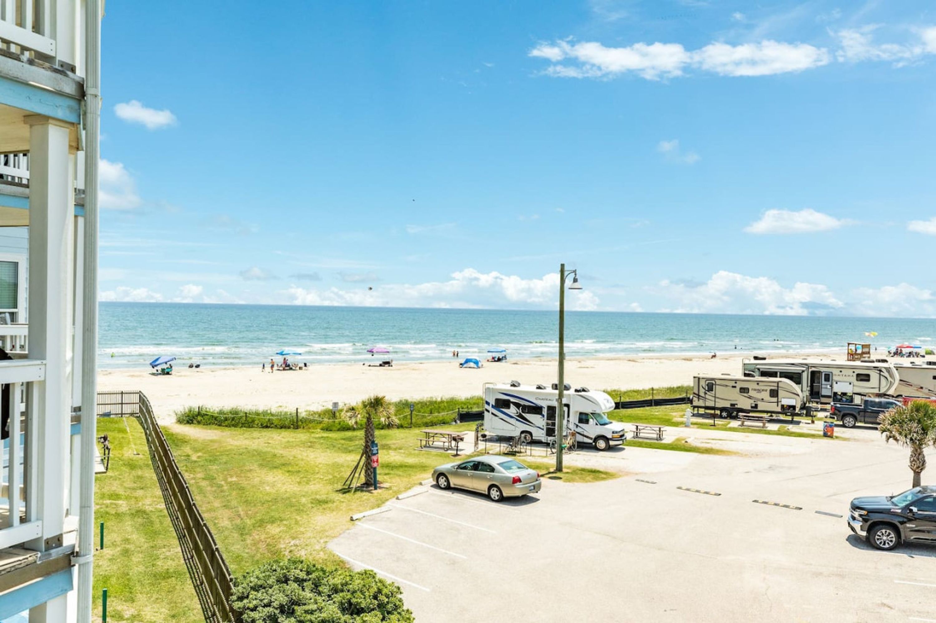 A beach view with RVs and cars parked, a parking lot, and a building with a balcony overlooking the beach.