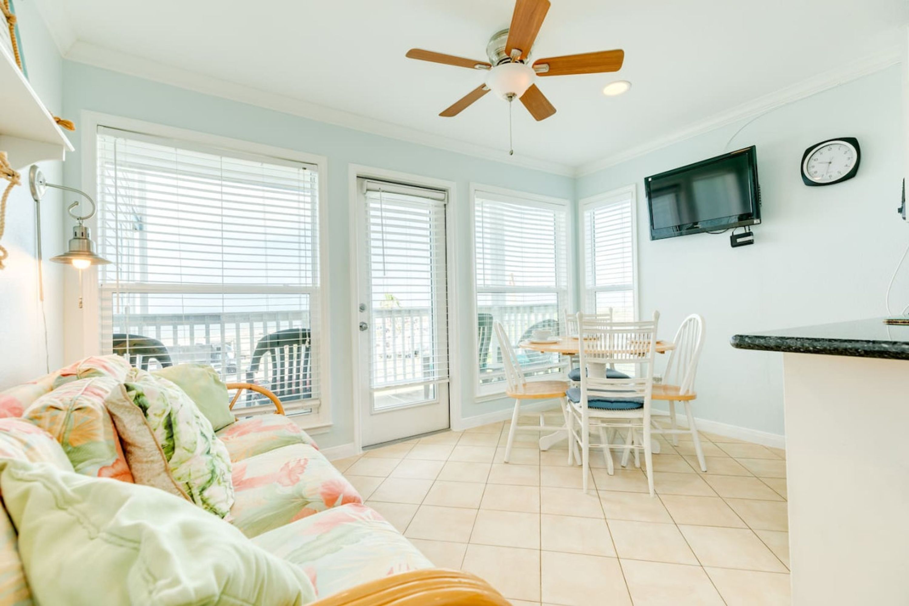 A bright room with a ceiling fan, a TV mounted on the wall, and a dining area with chairs and a table.