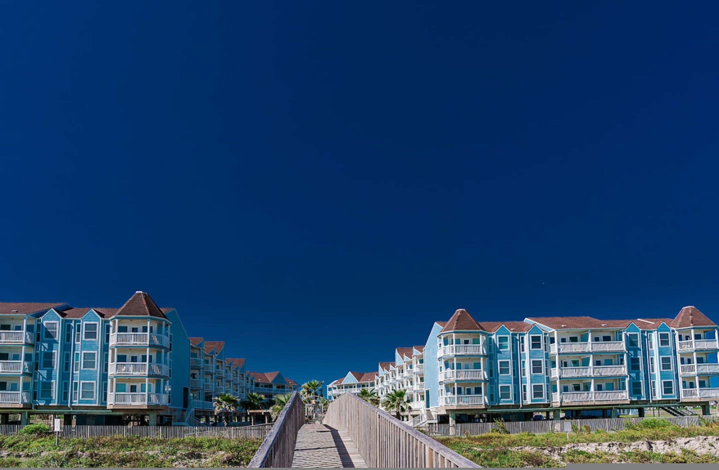 Una pasarela de madera que conduce a una hilera de edificios azules frente a la playa, con balcones.