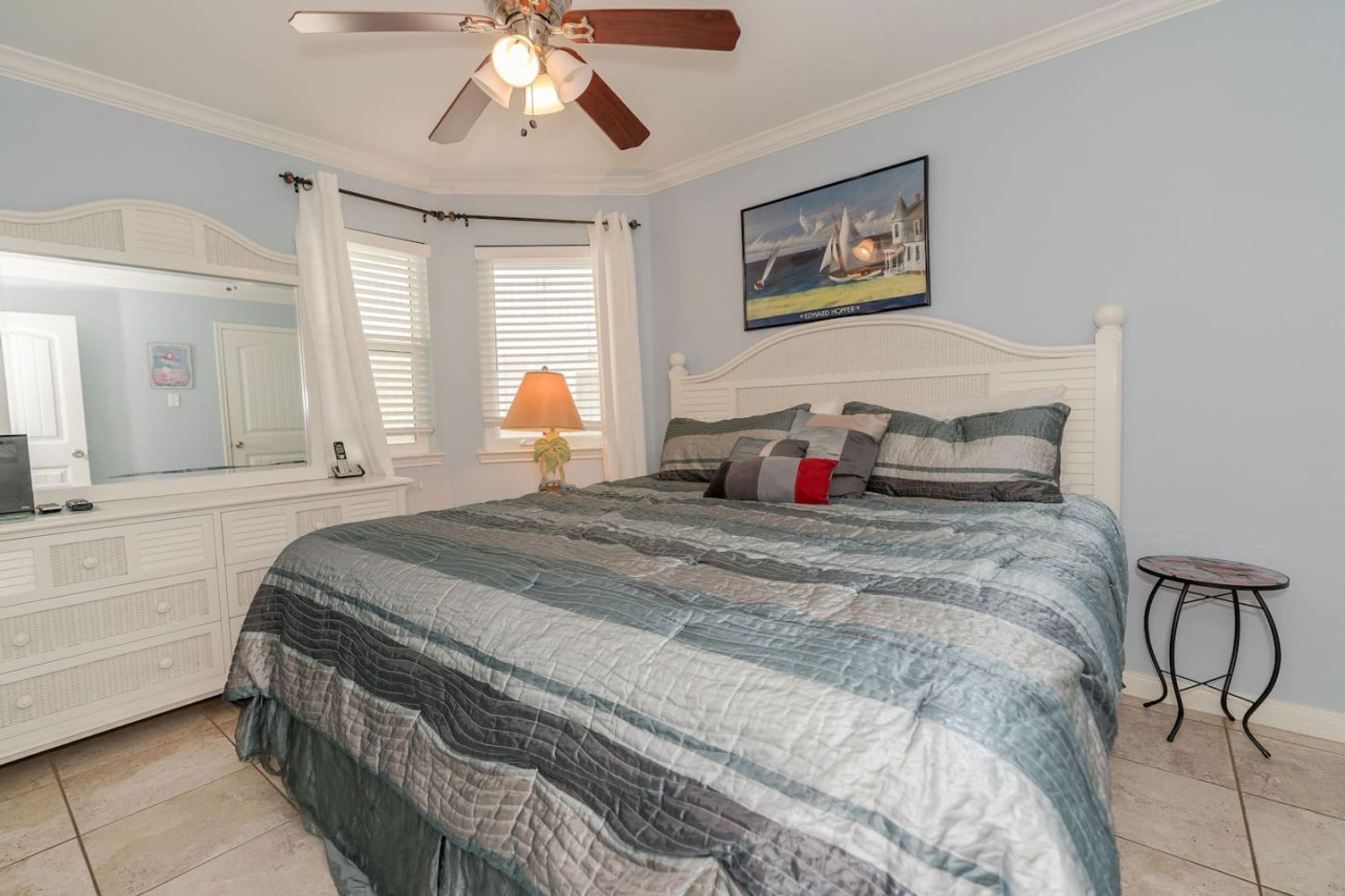 A bedroom with a bed, a ceiling fan, a nightstand, a lamp, a painting, and a window with curtains.