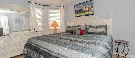 A bedroom with a bed, a ceiling fan, a nightstand, a lamp, a painting, and a window with curtains.