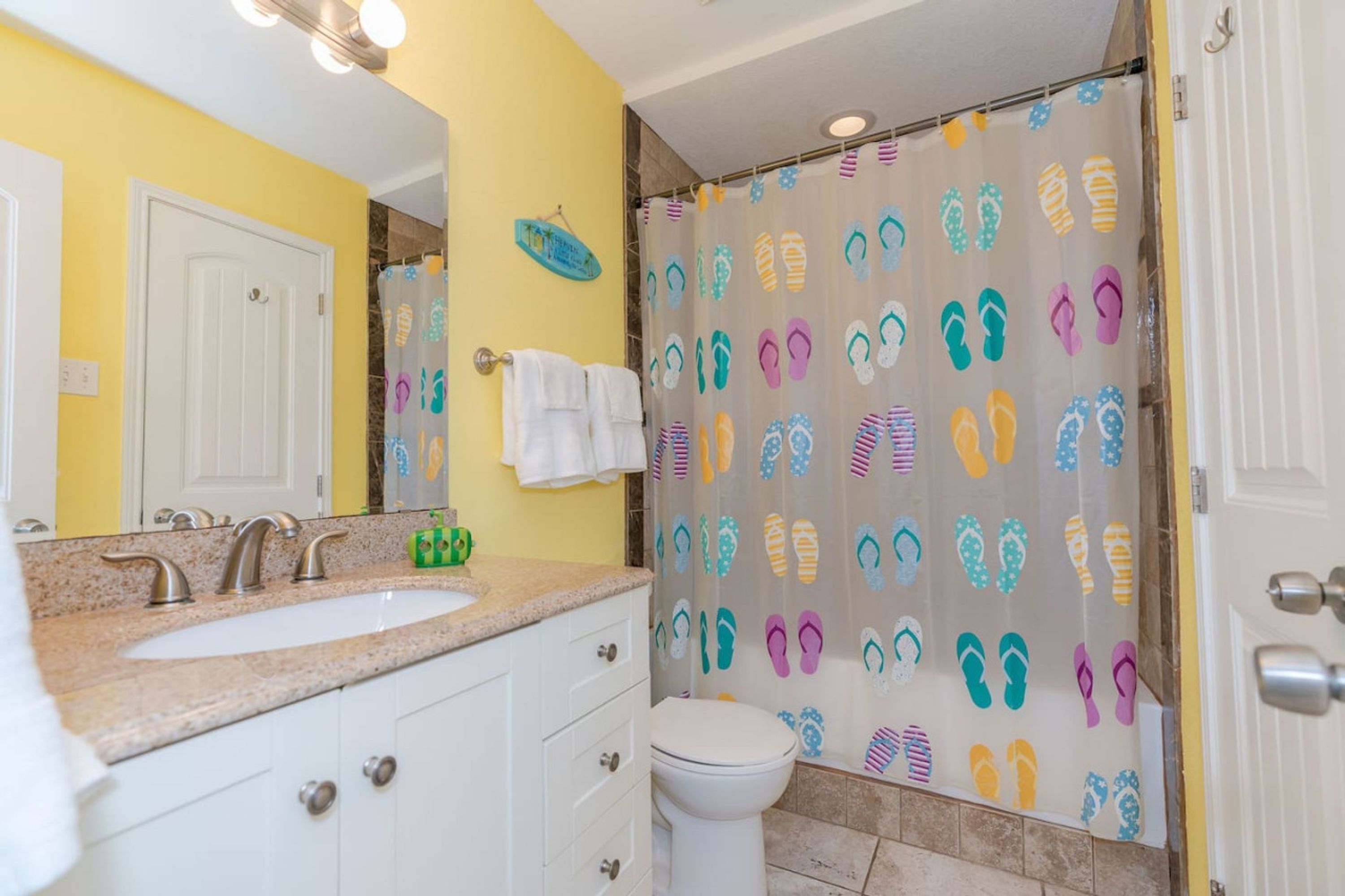 A bathroom with a yellow wall, a white sink, a toilet, and a shower curtain with a flip-flop pattern.