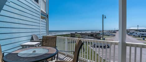 A covered patio with a table and chairs, overlooking a parking area and beach.
