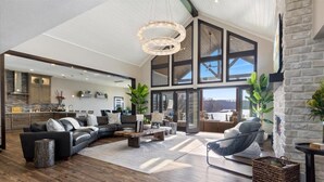A spacious living area with a large sectional sofa, a coffee table, and a fireplace. There is a kitchen in the background with a stone backsplash and wooden cabinets. - LOTO Pointe, Updated Luxury Home w/ Boat & PWC Slip, Boat Ramp, Firepits, Pool Table & More! (Versailles)