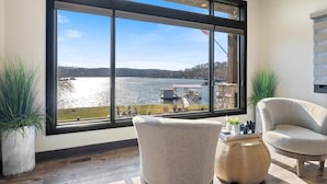 A room with a large window overlooking a lake, two chairs, a small table with decorative items, and a potted plant. - LOTO Pointe, Updated Luxury Home w/ Boat & PWC Slip, Boat Ramp, Firepits, Pool Table & More! (Versailles)