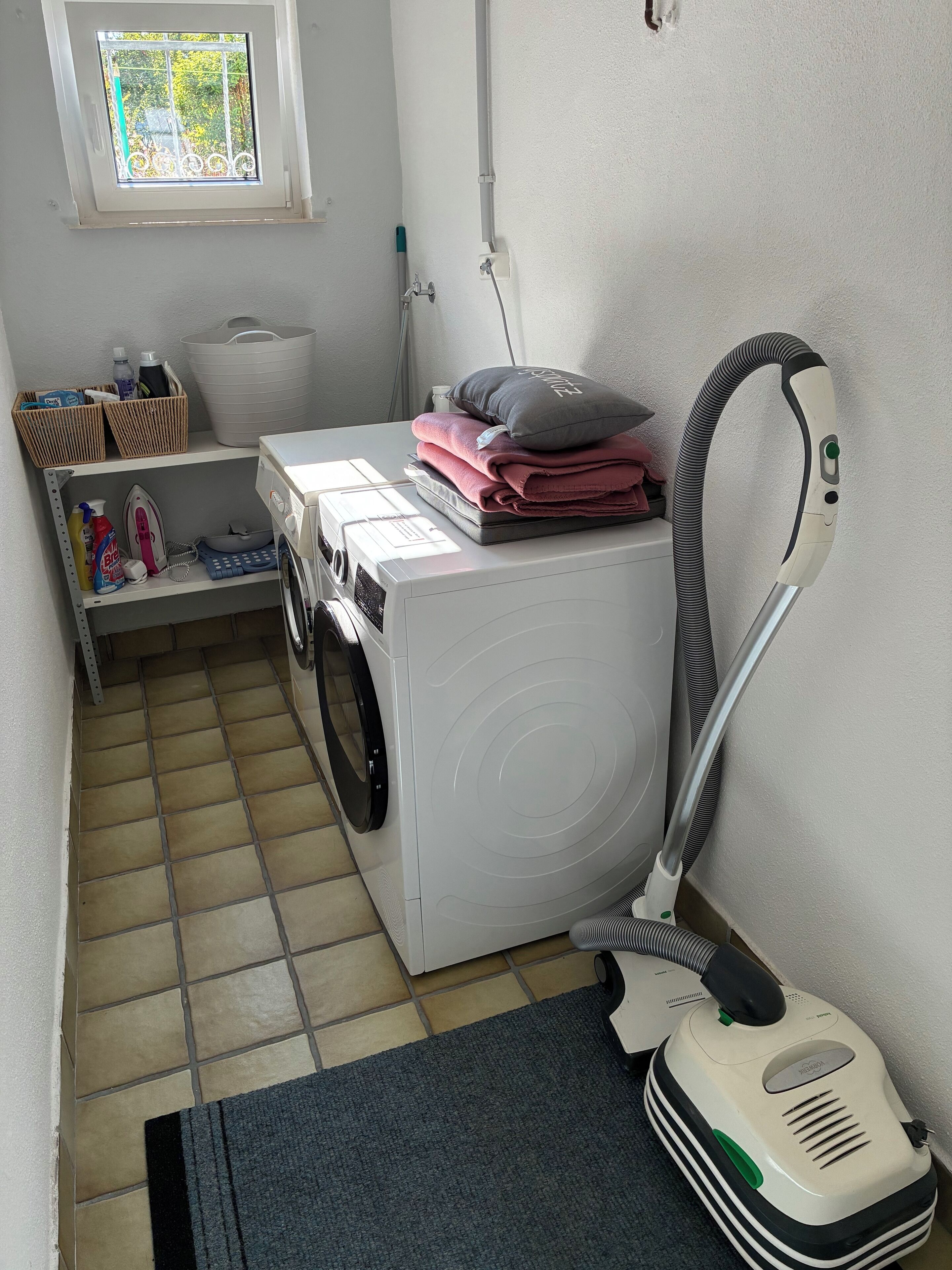 Un espace buanderie équipé d’un lave-linge, d’un aspirateur et d’une fenêtre donnant sur un paysage verdoyant.